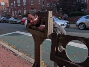 A playful moment at the Witch Dungeon Museum in Salem, Massachusetts, with a man leaning through a wooden stockade, surrounded by a backdrop of brick buildings and parked cars.