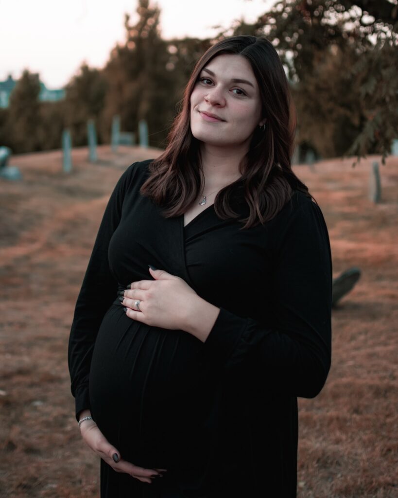 A serene image of a pregnant woman dressed in black, cradling her belly, with soft sunset lighting and a historic cemetery backdrop in Salem, Massachusetts.