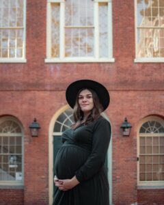 A pregnant woman in a black dress and hat stands in front of the historic Old Town Hall in Salem, Massachusetts, with its iconic red brick facade and arched windows.