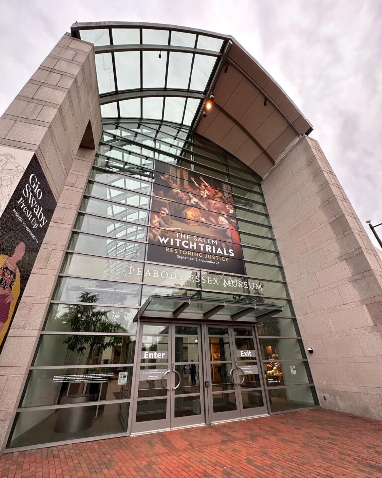 The tall glass facade of the Peabody Essex Museum in salem massachusetts, displaying a prominent banner for a witch trials exhibit above the main doors.