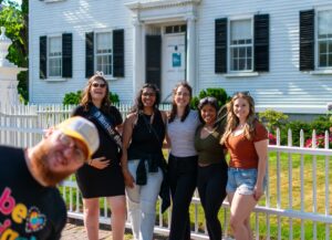 A group of friends posing in front of the Ropes Mansion in Salem, Massachusetts, with a man photobombing in the foreground, enjoying a sunny day.