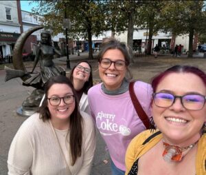A group of four smiling friends posing in front of the famous "Bewitched" statue in Salem, Massachusetts, surrounded by trees and a lively town square.