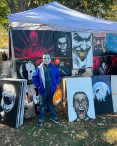 A person in a Michael Myers mask stands in front of a booth displaying vibrant horror-themed artwork, including characters like the Joker and Pennywise, at a Salem, Massachusetts fair.