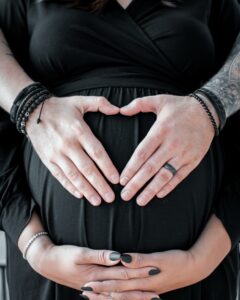 A close-up photo showing a pregnant belly with two sets of hands creating a heart shape, one set with black nail polish and the other with a ring and beaded bracelets, symbolizing love and anticipation.
