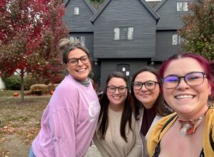 A group of friends smiling in front of the historic Witch House in Salem, Massachusetts, surrounded by vibrant autumn trees.