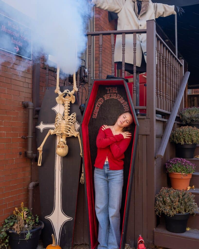 A woman posing inside a red coffin labeled "The Haunted Neighborhood," surrounded by Halloween decorations, including a skeleton and pumpkins, in Salem, Massachusetts.