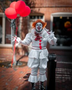 A detailed Pennywise cosplay in Salem, featuring the clown with vivid red balloons and a ruffled costume, posing menacingly on a brick street.