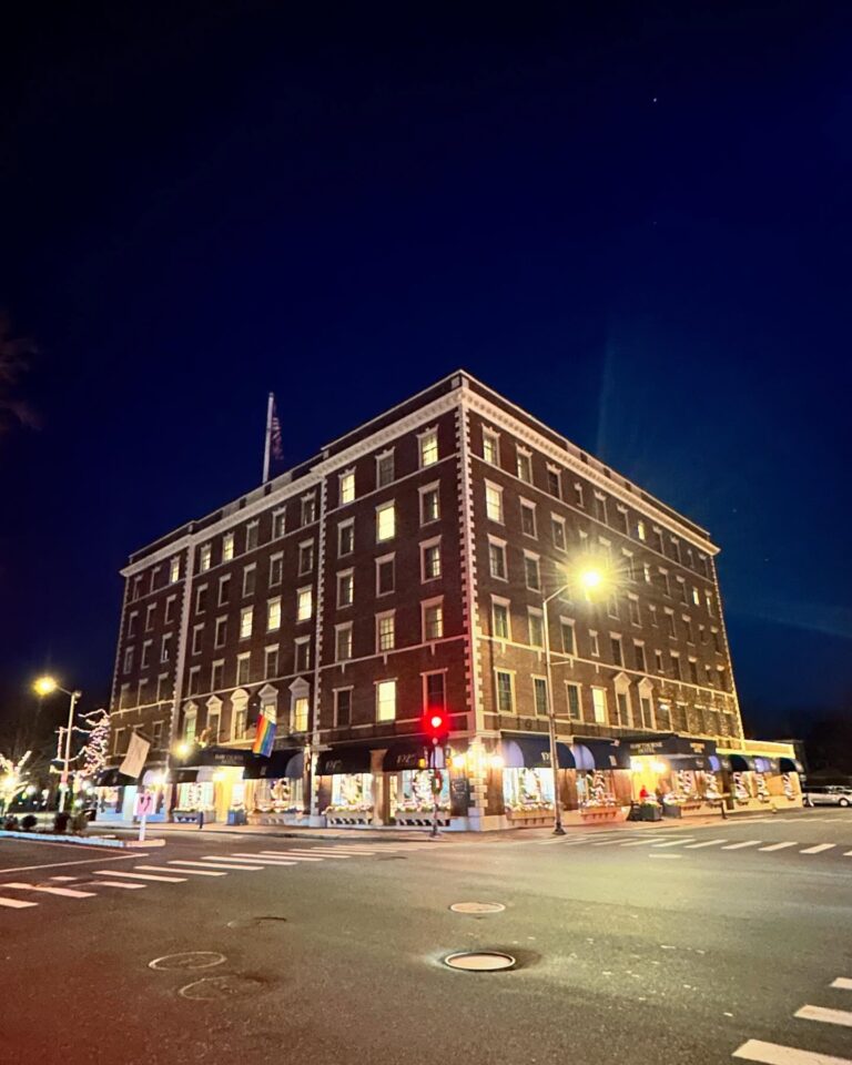 The Hawthorne Hotel in Salem, Massachusetts, lit up at night with glowing windows and festive decorations along the ground floor. The brick facade stands prominently against the dark blue sky.