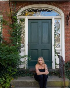 A young woman with blonde hair sits on the stone steps of a green door framed by ivy and a brick façade in Salem, Massachusetts, radiating calm and introspection.