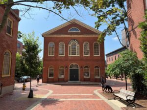 Salem's Old Town Hall, a red brick building with arched windows and a classic green door, surrounded by trees and cobblestone walkways, on a sunny day in Salem, Massachusetts.