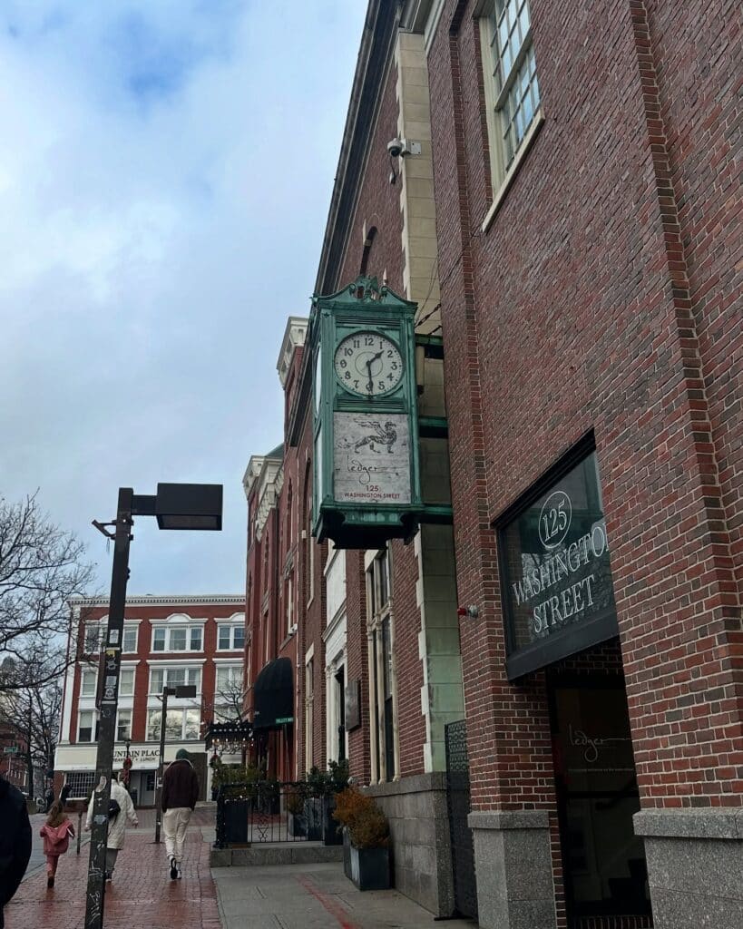 A green vintage clock hangs outside a brick building at 125 Washington Street in Salem, Massachusetts. The street is lined with red brick buildings, black awnings, and pedestrians.