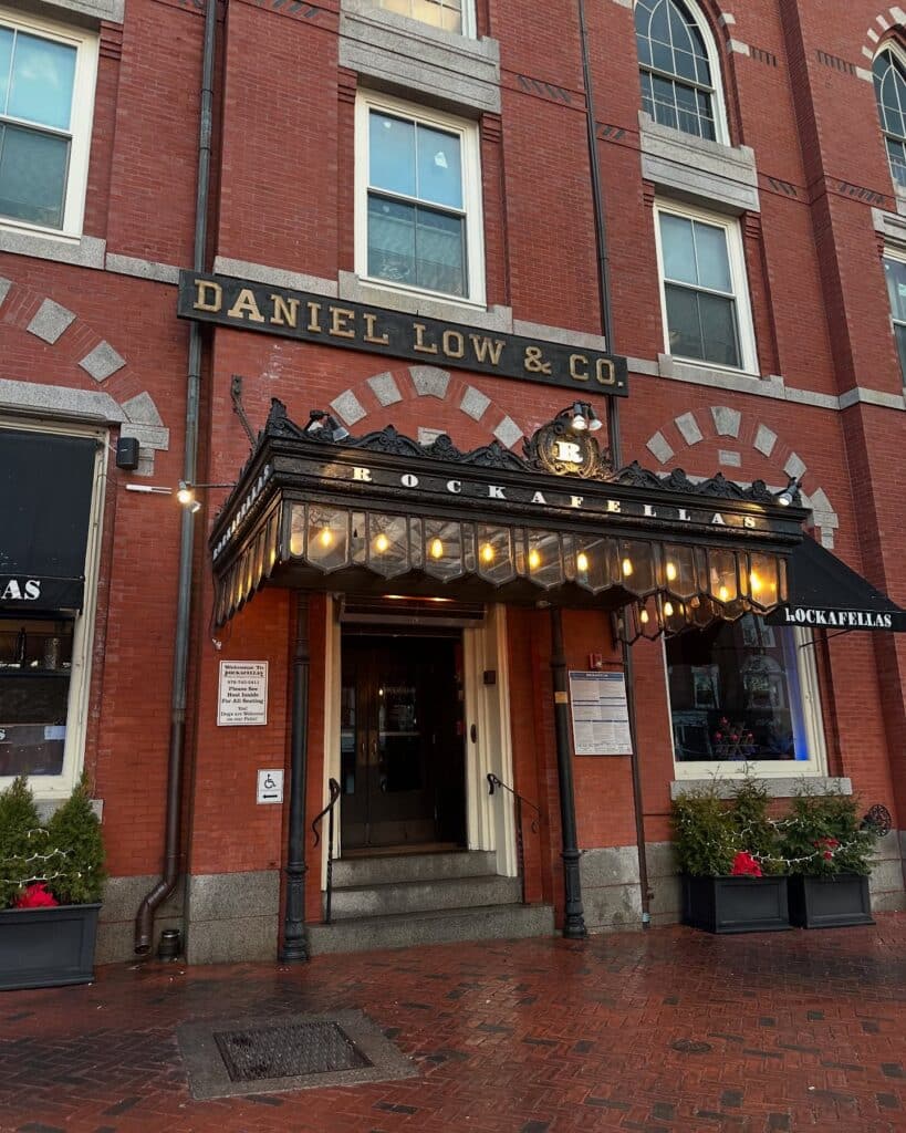 The entrance to Rockafellas restaurant in Salem, Massachusetts, located in the historic Daniel Low & Co. building with brick walls, large windows, and an elegant black canopy with glowing lights.