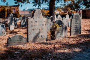 A row of historic gravestones at the Old Burying Point Cemetery in Salem, Massachusetts, with autumn leaves scattered on the ground and a mix of trees and urban buildings in the background.