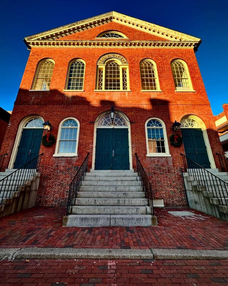 The Old Town Hall in Salem, Massachusetts, a two-story red brick building with arched windows and green double doors, bathed in golden sunlight with holiday wreaths on the doors.