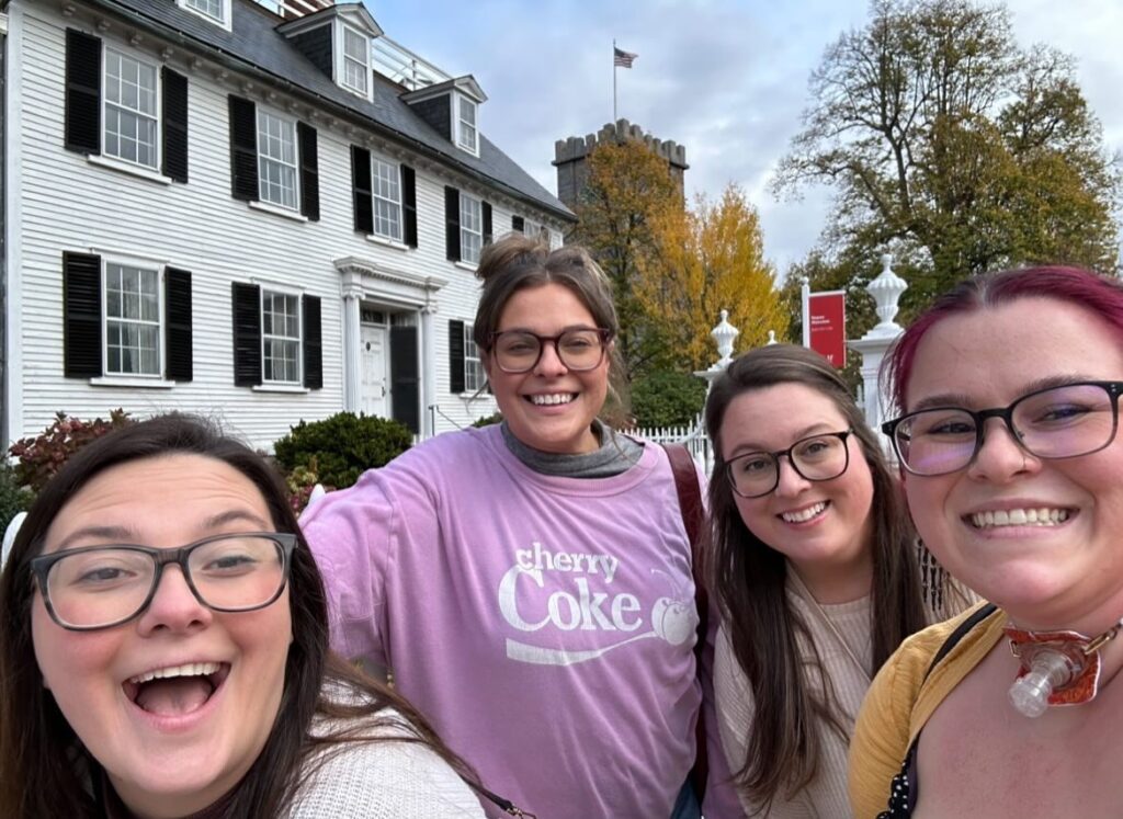 A group of four friends poses happily in front of the historic white Ropes Mansion with black shutters in Salem, Massachusetts, surrounded by autumn trees and a bright, cheerful atmosphere.