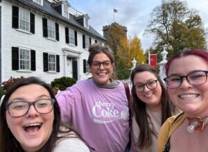 A group of four friends poses happily in front of the historic white Ropes Mansion with black shutters in Salem, Massachusetts, surrounded by autumn trees and a bright, cheerful atmosphere.