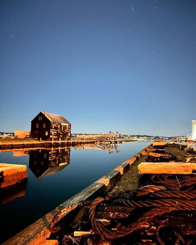 The Salem Sail Loft is reflected in the calm harbor waters under a clear, starry night sky. The foreground features weathered ropes and dock materials, adding to the maritime atmosphere