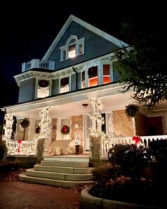 A beautifully decorated house in Salem, Massachusetts, illuminated at night with warm white string lights, red bows, and holiday wreaths on the porch and windows.
