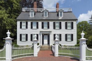 The Ropes Mansion in Salem, Massachusetts, a historic white colonial-style home with black shutters, brick chimneys, and a charming white picket fence under a bright blue sky.