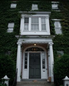 The front door of a historic ivy-covered mansion in Salem, Massachusetts, featuring white columns, intricate window designs, and an inviting green door.