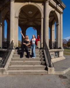 Two women enjoying a sunny day at the Salem Common Bandstand in Salem, Massachusetts. They are posing happily on the steps of the historic structure, framed by its elegant arches and the American flag in the background.