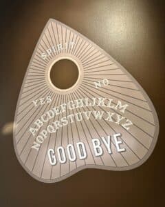 A large Ouija board planchette-shaped wall decoration in Salem, Massachusetts, featuring letters, "Yes," "No," "Spirit," and "Good Bye" written on it. The design is light with bold white text.