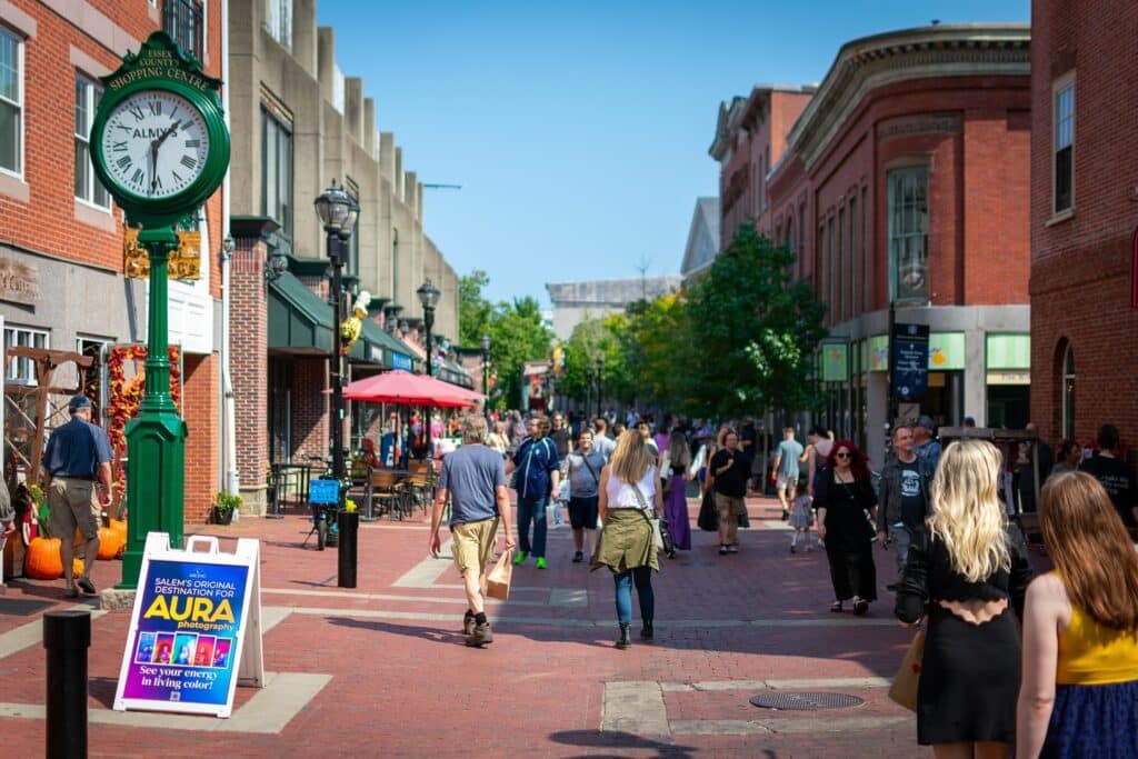 A bustling scene on Essex Street in Salem, Massachusetts, featuring people walking along the pedestrian-friendly brick pathway surrounded by shops, cafes, and historic buildings.