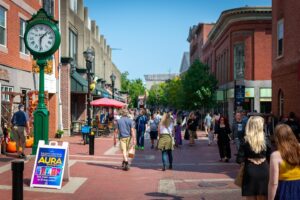 A bustling scene on Essex Street in Salem, Massachusetts, featuring people walking along the pedestrian-friendly brick pathway surrounded by shops, cafes, and historic buildings.