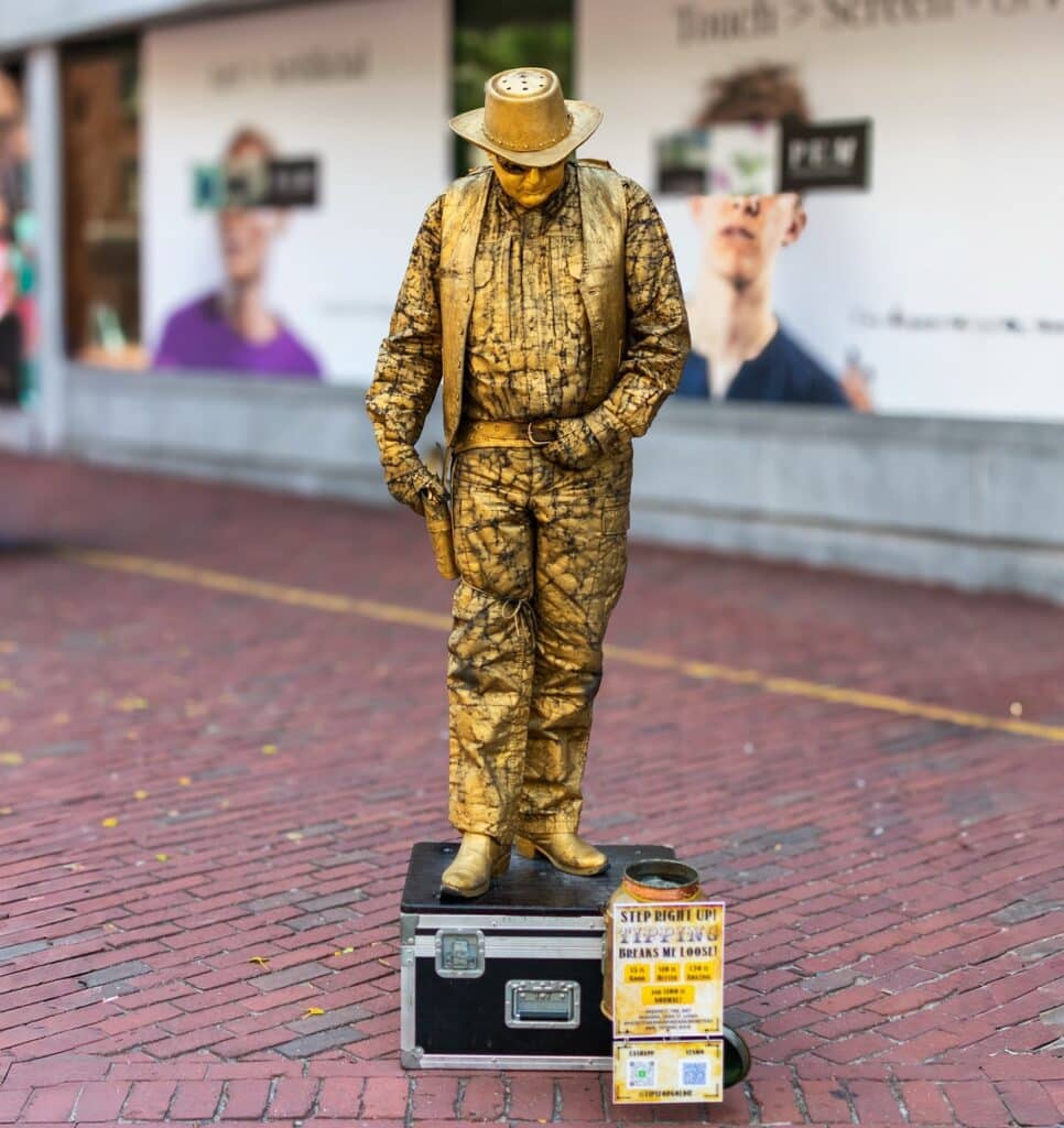 A gold-painted living statue dressed in cowboy attire, standing motionless on a black platform along the red brick pavement of Essex Street in Salem, Massachusetts.