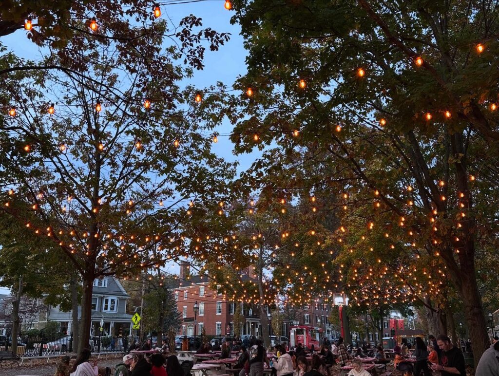 Twinkling string lights illuminate the trees over Salem Common in Salem, Massachusetts, as people gather under the cozy glow at dusk.