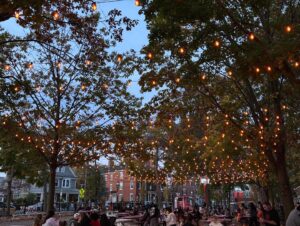 Twinkling string lights illuminate the trees over Salem Common in Salem, Massachusetts, as people gather under the cozy glow at dusk.