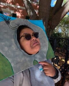 A person in Salem, Massachusetts, playfully making a silly face while holding up a quirky, hand-painted frame outdoors with trees and autumn vibes in the background.