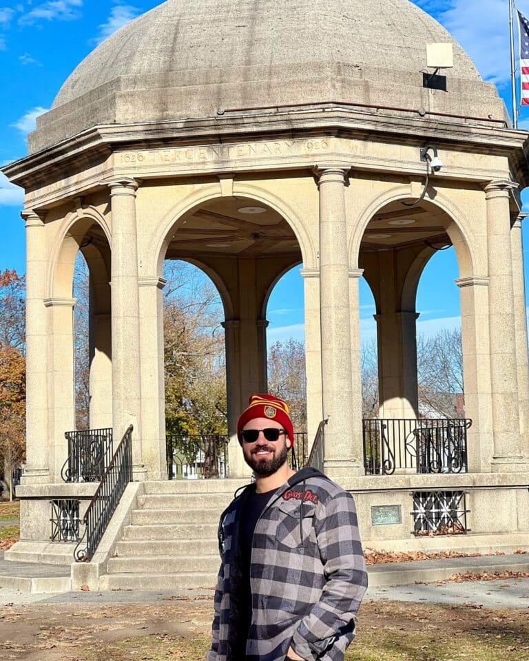 A man in sunglasses and a flannel stands in front of the Salem Common Bandstand in Salem, Massachusetts. The stone structure features classic columns and a domed roof with "Tercentenary 1626-1926" engraved on it.