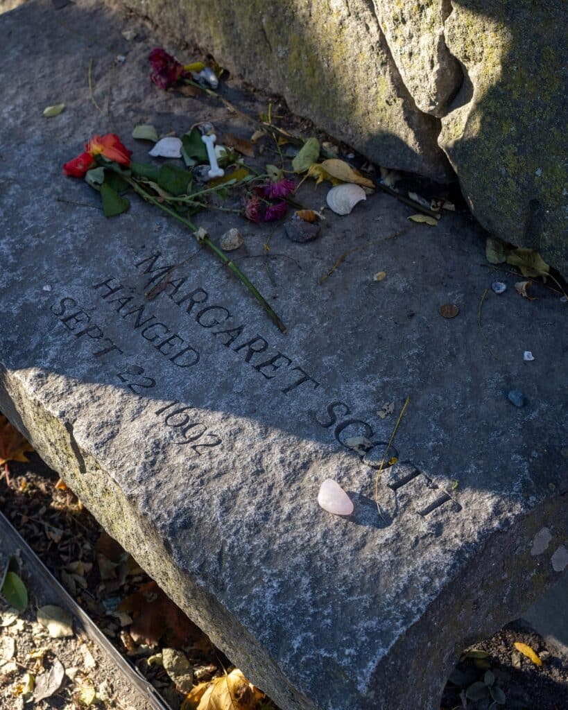 A stone memorial bench dedicated to Margaret Scott, one of the victims of the Salem Witch Trials, engraved with the date "Sept. 22, 1692." Flowers, coins, and small tokens are left as tributes at the Salem Witch Trials Memorial in Salem, Massachusetts.