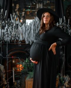 A pregnant woman in a black dress and wide-brimmed hat stands confidently in a candlelit gothic setting in Salem, Massachusetts, surrounded by cascading wax candles and moody decor.