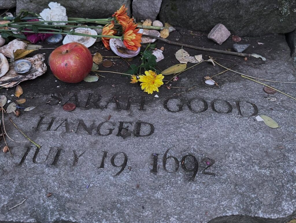 A memorial stone for Sarah Good, inscribed with "Hanged July 19, 1692," at the Salem Witch Trials Memorial in Salem, Massachusetts, adorned with flowers, coins, an apple, and seashells.