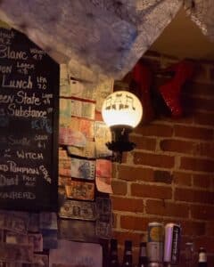 A bar corner inside Mercy Tavern in Salem Massachusetts featuring a glowing "BAR" light, red shoes on a brick wall, and international currency pinned beside a chalkboard drink menu.