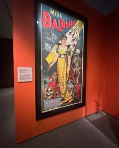 A vintage poster of Miss Baldwin, "A Modern Witch of Endor," displayed at the Peabody Essex Museum in Salem, Massachusetts. The artwork shows Miss Baldwin surrounded by skeletons, demons, and ghostly figures.