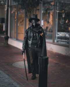 A person dressed as a plague doctor in a leather coat, black hat, and mask, holding a cane while walking on a brick sidewalk in Salem, Massachusetts.