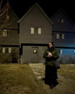 A woman stands at night in front of the Witch House in Salem, Massachusetts, holding a candle. The historic black house with leaded glass windows and a wreath on the door is dimly lit in the background.