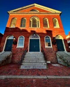 The Old Town Hall in Salem, Massachusetts, stands tall with its red brick facade, green double doors, and arched windows, illuminated by the warm evening sun. Festive wreaths hang by the entrance during the holiday season.