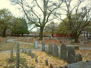 The Old Burying Point Cemetery in Salem, Massachusetts, with historic gravestones surrounded by fallen leaves, bare trees, and a quiet, reflective atmosphere.