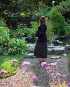 A serene view of a pregnant woman in a flowing black dress walking through the vibrant Ropes Mansion Garden in Salem, Massachusetts, surrounded by colorful flowers, greenery, and a small pond.