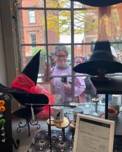 A woman in a pink sweater smiles and waves through a window of a Salem, Massachusetts shop filled with witch-themed items like hats, mugs, and zodiac mystery crystal boxes.