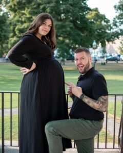 A playful image of a pregnant woman posing confidently while her partner kneels beside her, humorously pointing at her belly, set in the open green space of Salem Common in Salem, Massachusetts.