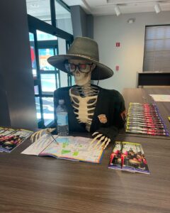 A skeleton wearing a park ranger hat and glasses sits at a table in Salem, Massachusetts, with a map, brochures, and colorful pens, creating a fun and spooky scene.