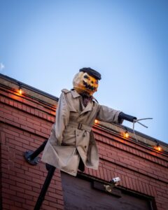 A pumpkin-headed scarecrow dressed in a trench coat and beret, with claw-like hands, leans over a rooftop in Salem, Massachusetts, surrounded by glowing orange string lights.