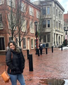 A woman in a black winter coat and orange purse stands on Essex Street in Salem, Massachusetts, surrounded by brick buildings, bare trees, and holiday decorations.