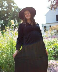 A joyful pregnant woman in a black dress and wide-brimmed hat smiling brightly in the sunlit Ropes Mansion Garden, surrounded by colorful flowers in Salem, Massachusetts.