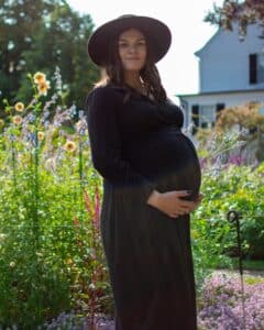 A pregnant woman in a black dress and wide-brimmed hat cradles her belly while standing in the Ropes Mansion Garden in Salem, Massachusetts, surrounded by blooming flowers and greenery.